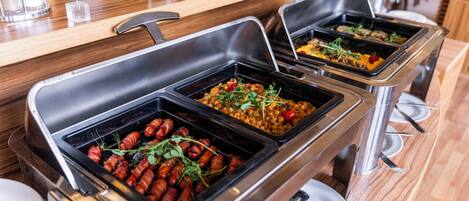 Buffet-style food service with various dishes, including bacon-wrapped items and salads, in a modern setting with wooden tables and metal serving trays.