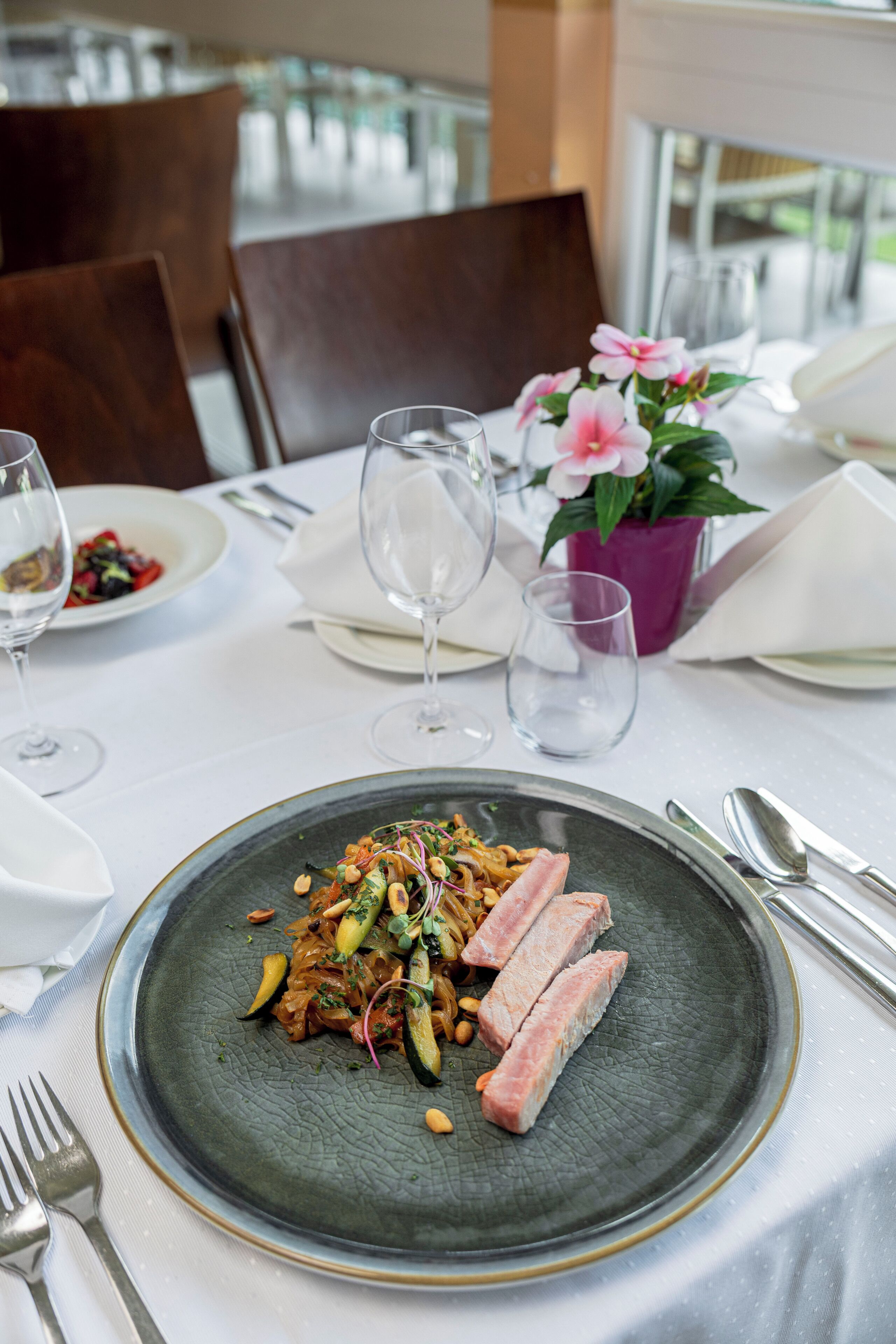 Una mesa de comedor dispuesta con platos, cubiertos y vasos, con un centro de mesa floral y una silla al fondo.