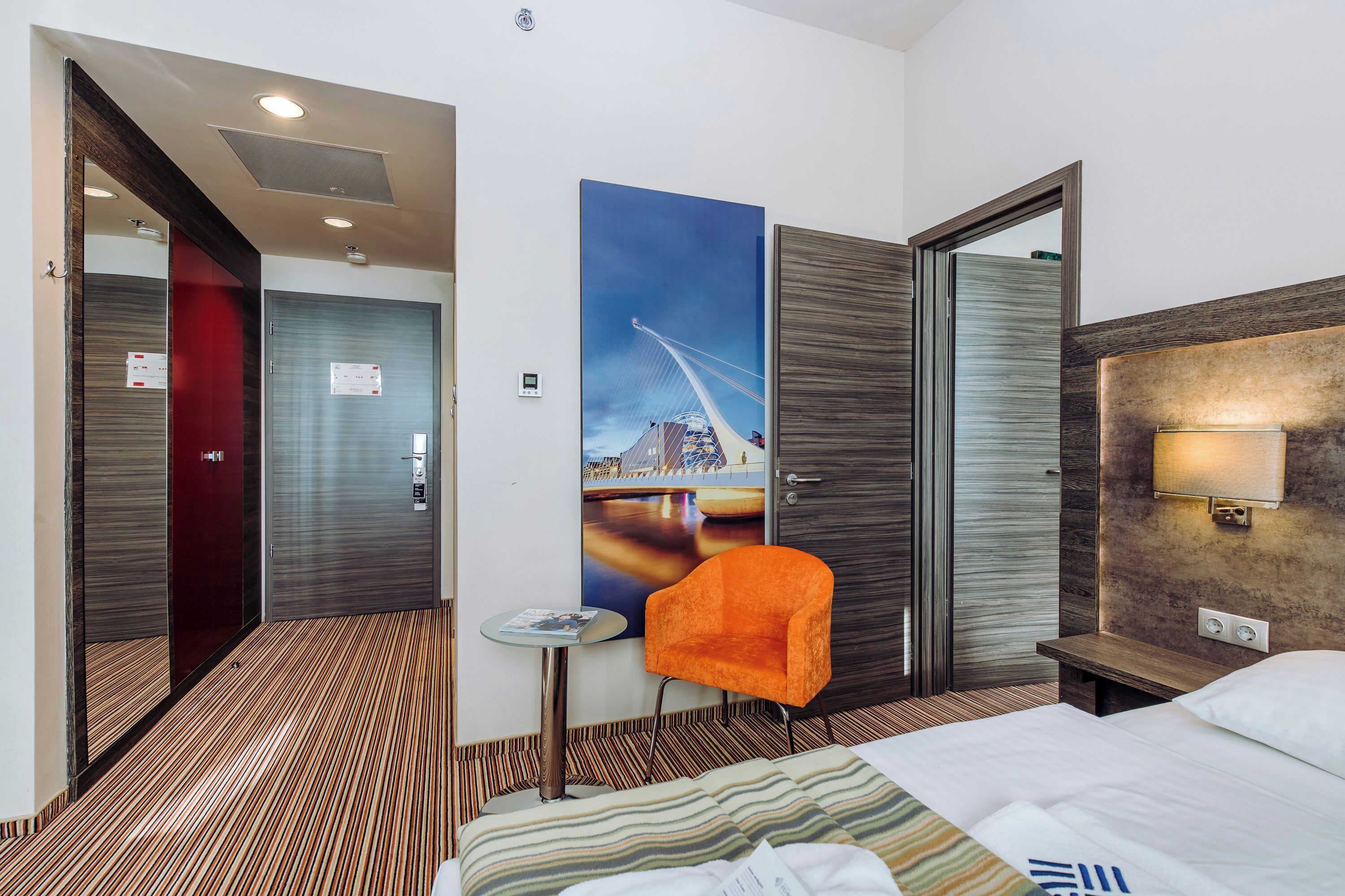Una habitación de hotel con una cama, una silla, un cuadro de un puente y un cabecero de madera.