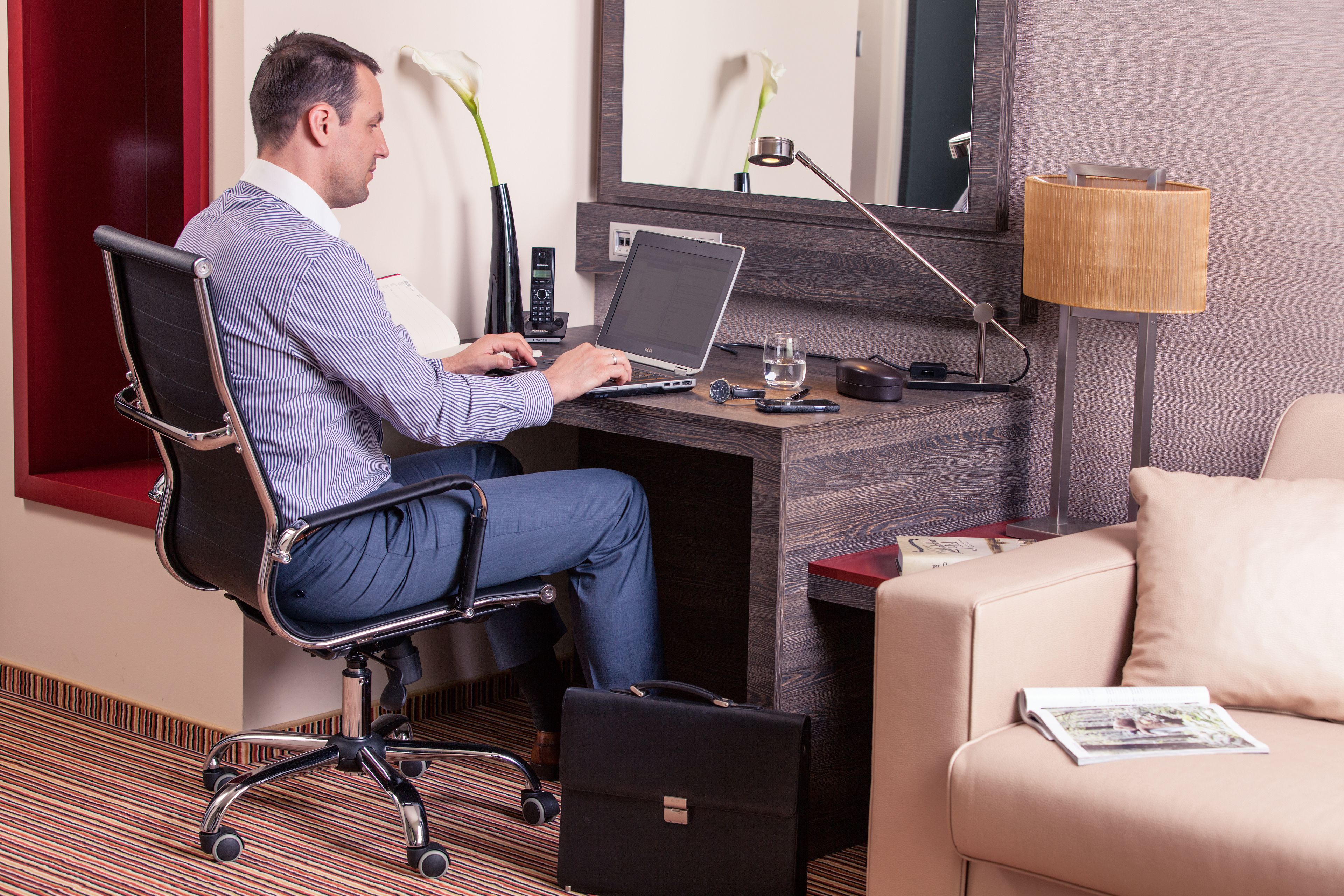 A man working at a desk in a hotel room with a laptop, a desk lamp, a glass of water, a magazine, and a briefcase.