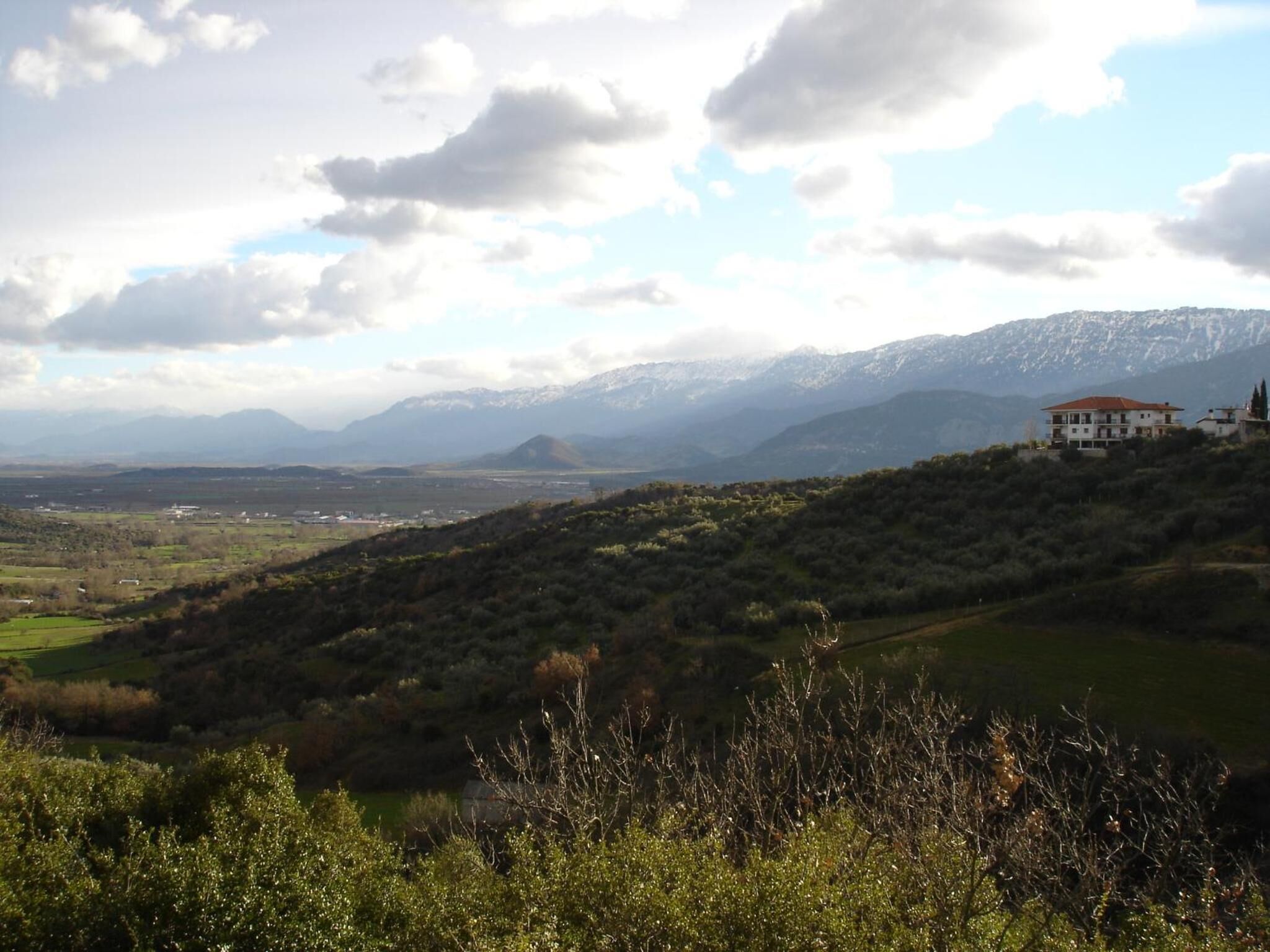 A mountainous landscape with a house on a hill, surrounded by greenery and distant mountains.