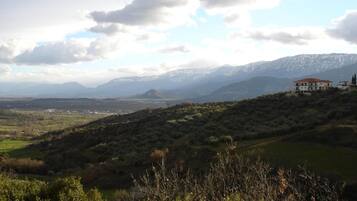 A mountainous landscape with a house on a hill, surrounded by greenery and distant mountains.