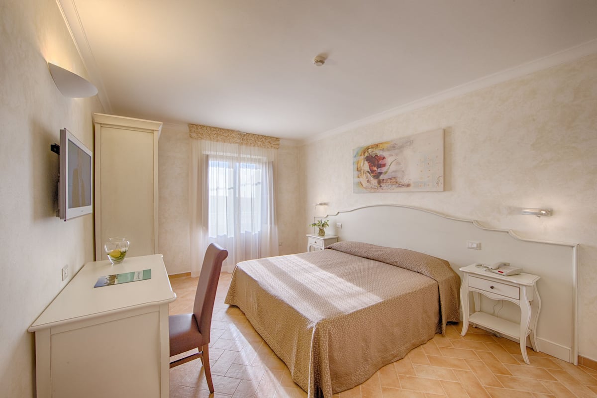 A hotel room with a bed, a desk, a chair, a bedside table, and a painting on the wall.