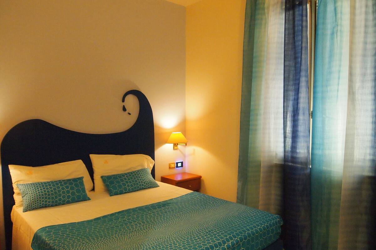 Una cama bien hecha con una colcha estampada, una lámpara de pared y una ventana con cortinas.