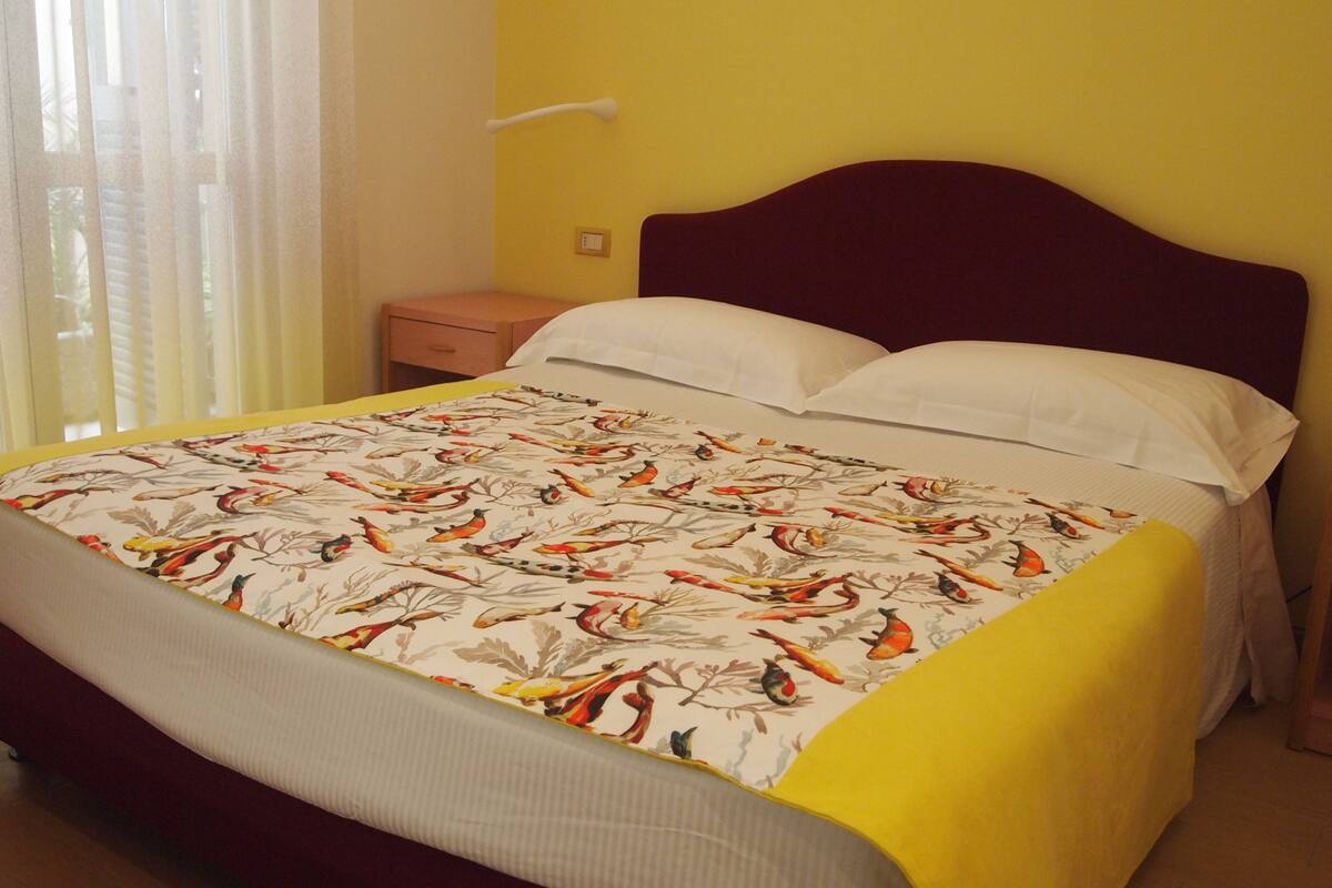 Una cama con una colcha estampada con motivos florales, una mesita de noche de madera y una mesita con una lámpara.