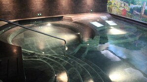 Una piscina con fondo de cristal y baranda de metal, rodeada de muros de piedra y un mural de un pueblo.