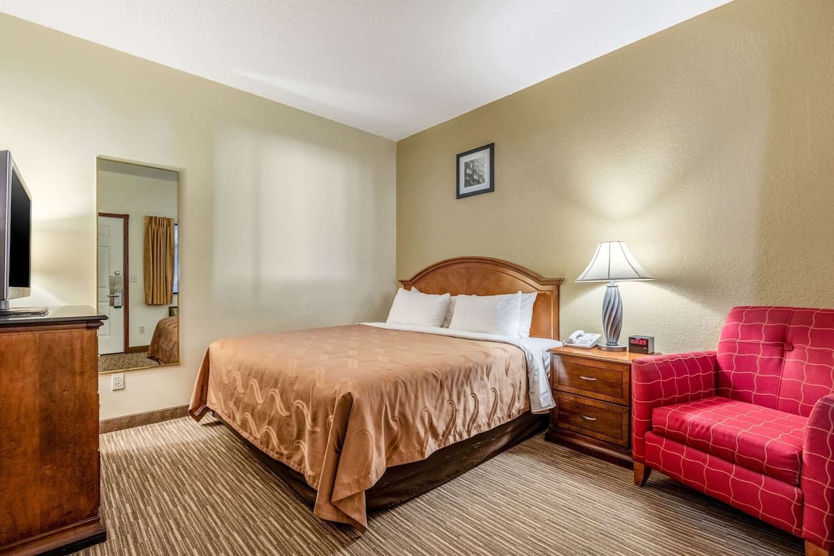 A hotel room with a bed, a desk, a chair, a lamp, and a television.