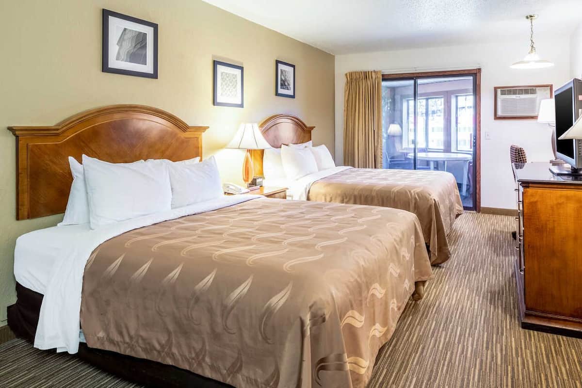 A hotel room with two beds, a desk with a computer, a chair, and a view of the outdoors through a sliding door.
