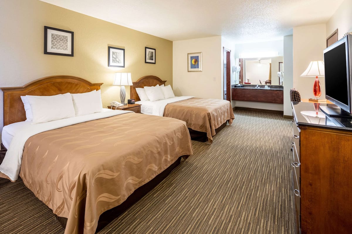 A hotel room with two beds, a TV, a desk, and a bathroom visible through an open door.