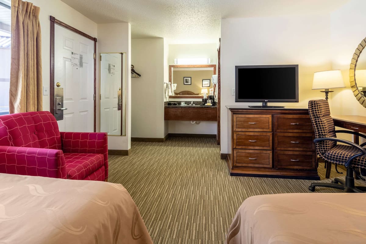 A hotel room with two beds, a television, a desk with a chair, a dresser, and a bathroom visible in the background.
