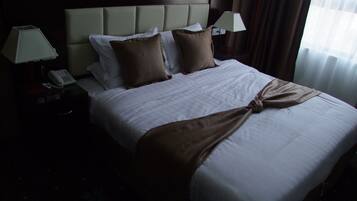 A neatly made bed with a brown throw, two pillows, and a headboard. There is a bedside table with a lamp and a phone.