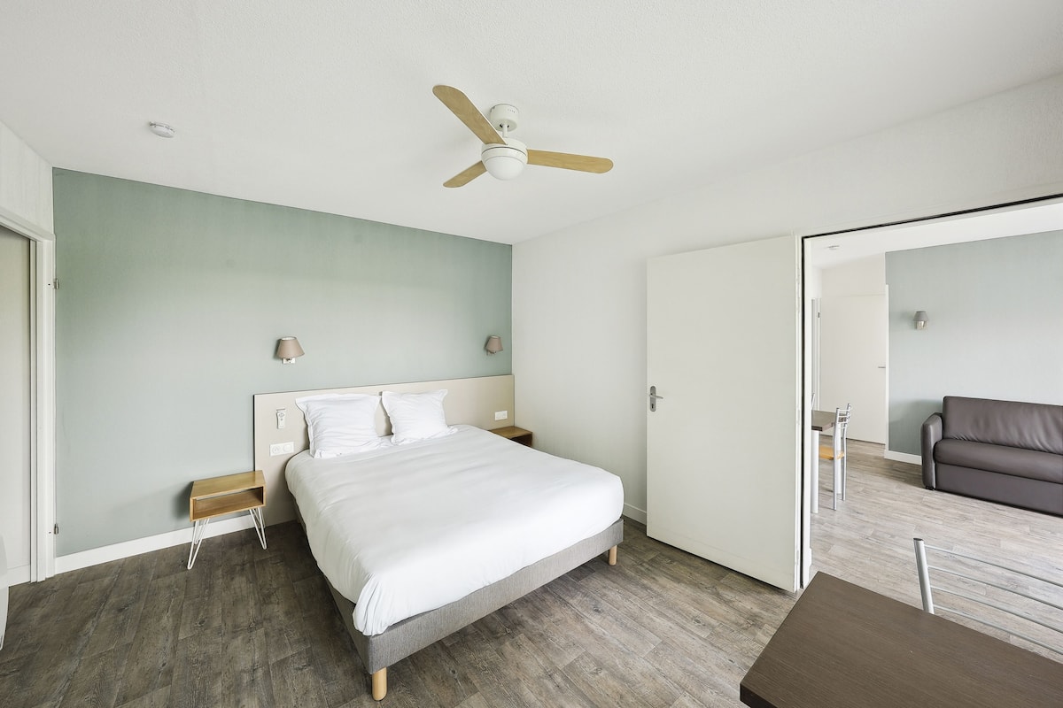 A bedroom with a bed, a ceiling fan, a nightstand, a bedside table, and a door leading to another room.