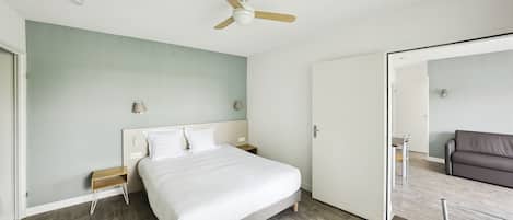 A bedroom with a bed, a ceiling fan, a nightstand, a bedside table, and a door leading to another room.