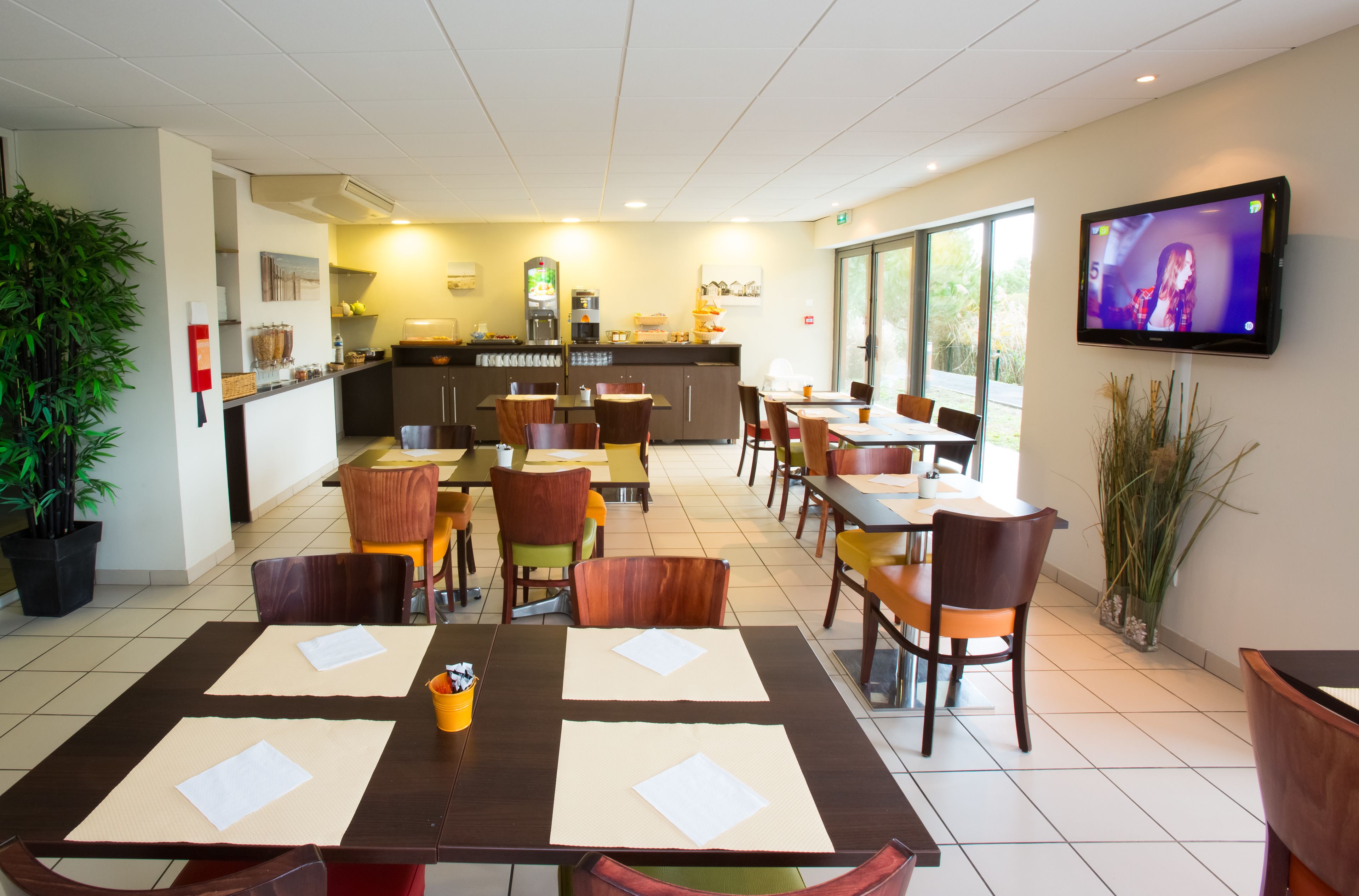 A restaurant with multiple tables, chairs, a kitchen area, and a TV screen.