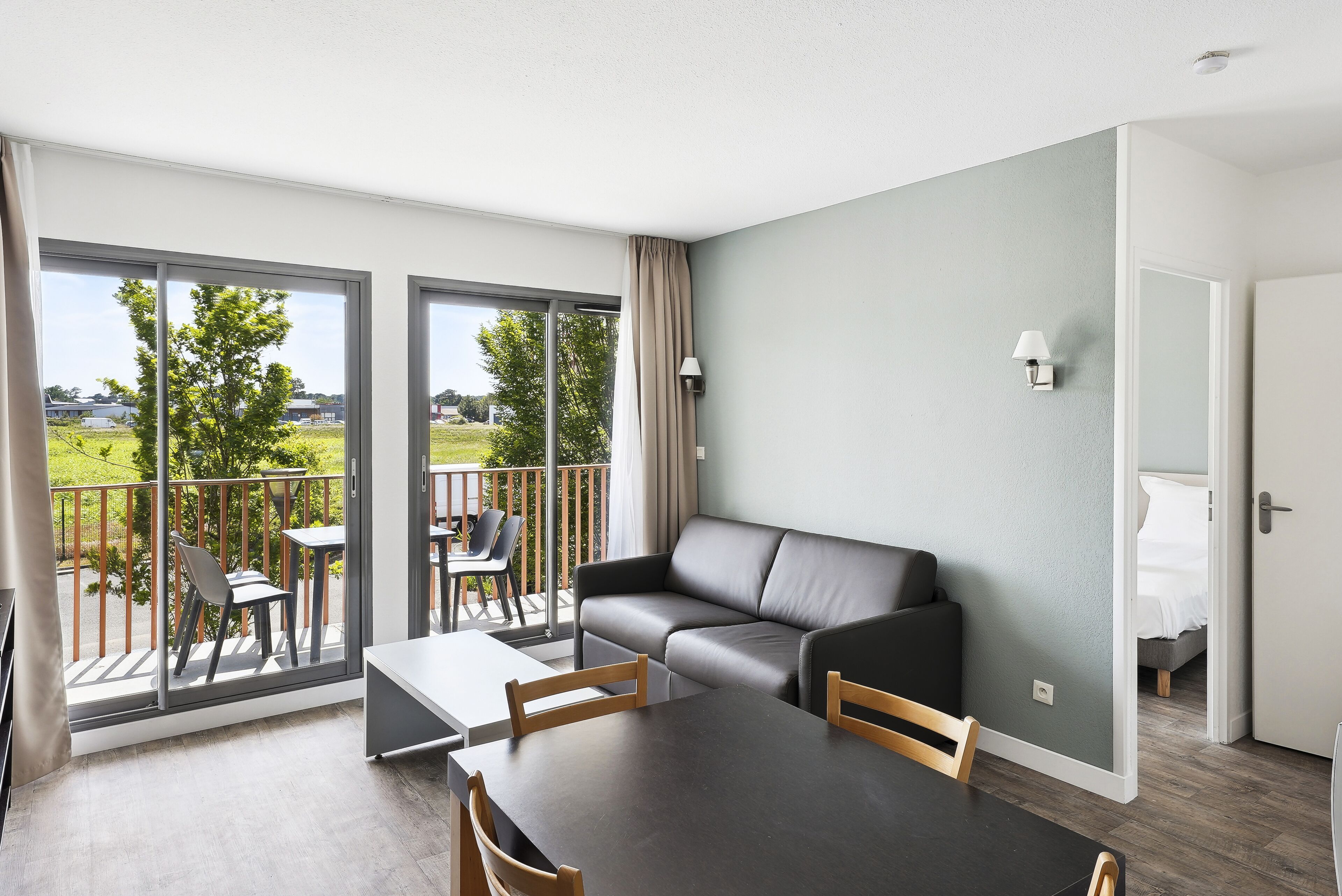 Ein modernes Wohnzimmer mit einer Couch, einem Esstisch und Stühlen, ein Balkon mit Blick und eine Tür zu einem Schlafzimmer.