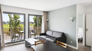Una sala de estar moderna con sofá, mesa de comedor y sillas, un balcón con vistas, y una puerta que da a un dormitorio.
