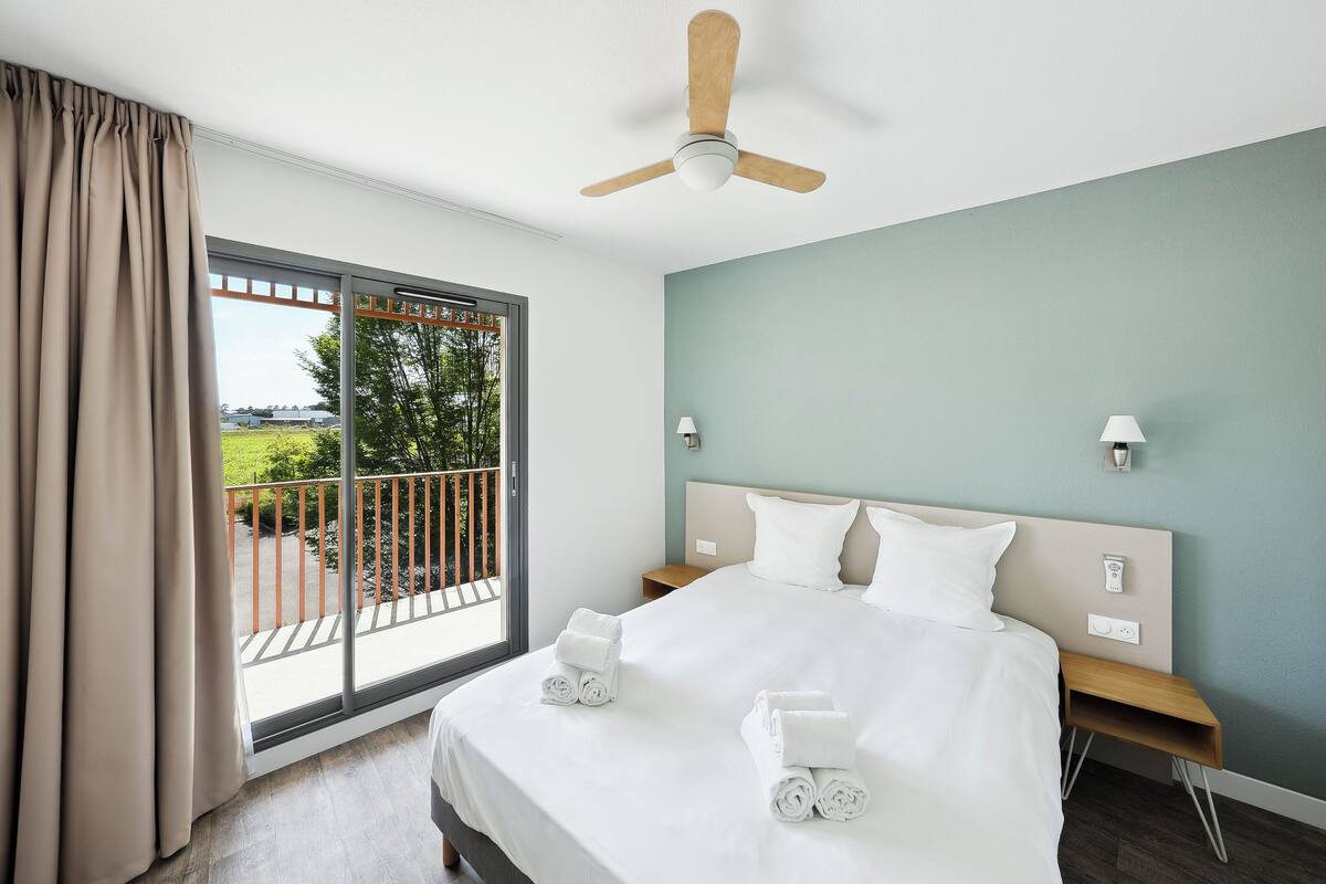 A hotel room with a large bed, a ceiling fan, a nightstand, a lamp, and a view of a balcony with a railing.