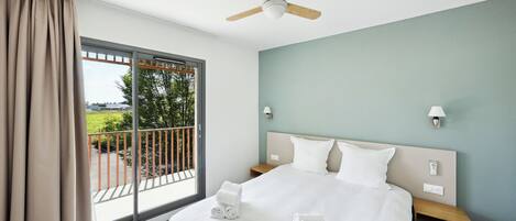 A hotel room with a large bed, a ceiling fan, a nightstand, a lamp, and a view of a balcony with a railing.