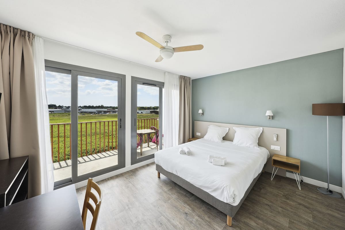 A hotel room with a large bed, a desk, a chair, a balcony with a view of the countryside, and a ceiling fan.