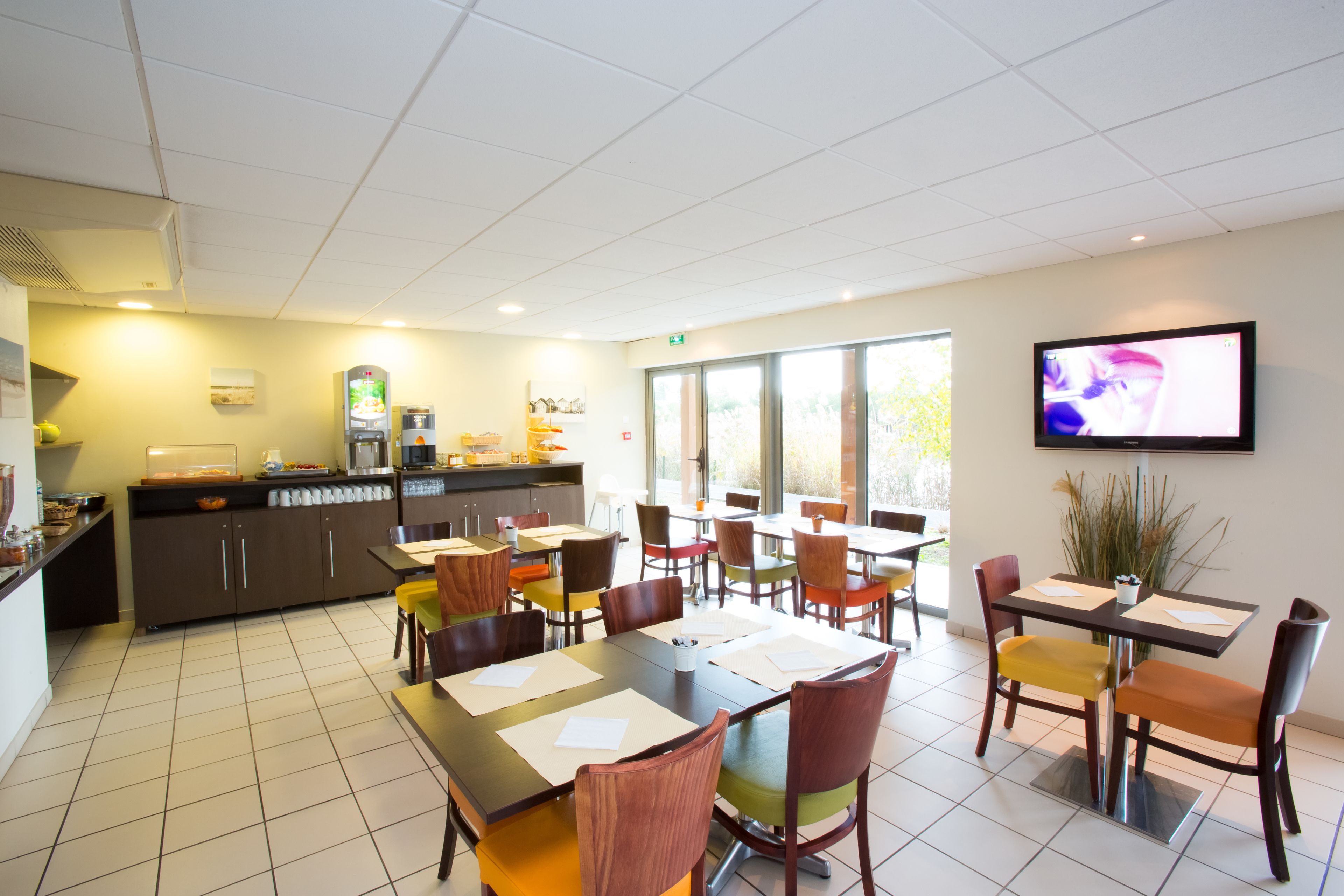 A dining area with multiple tables and chairs, a kitchen area with a refrigerator, and a television mounted on the wall.