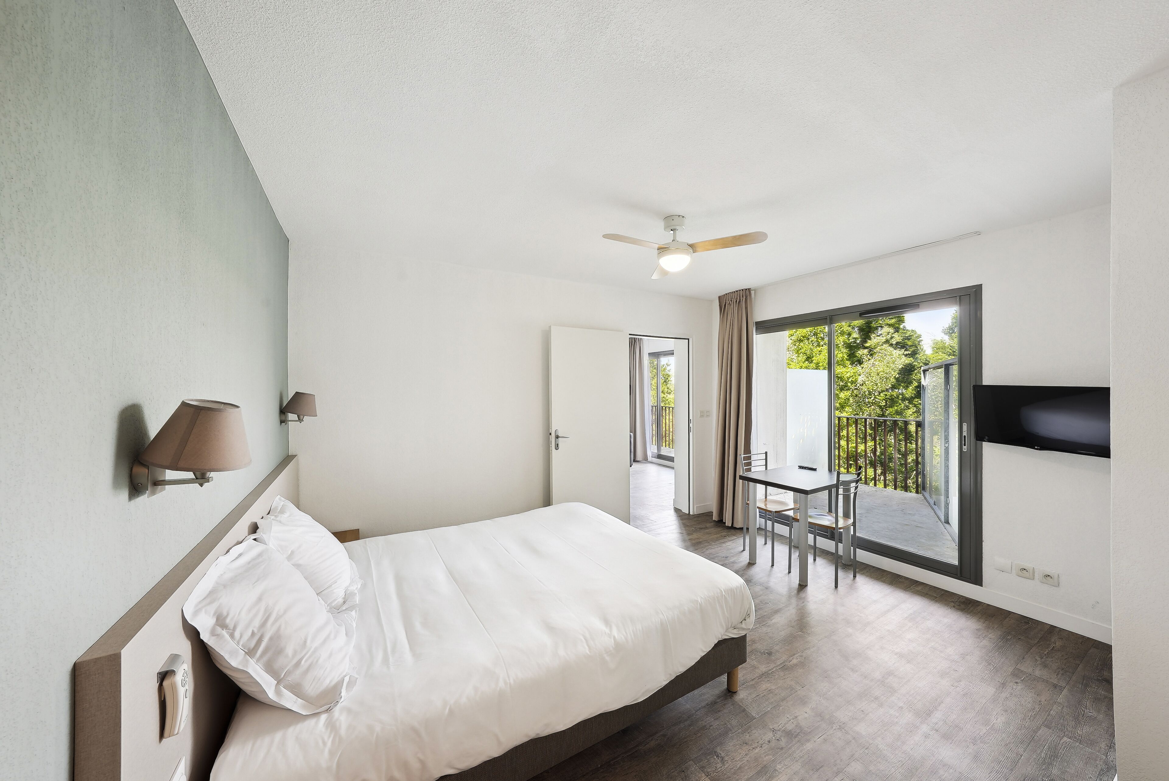 Een slaapkamer met een bed, een plafondventilator, een aan de muur bevestigde televisie en een balkon met een tafel en stoelen.