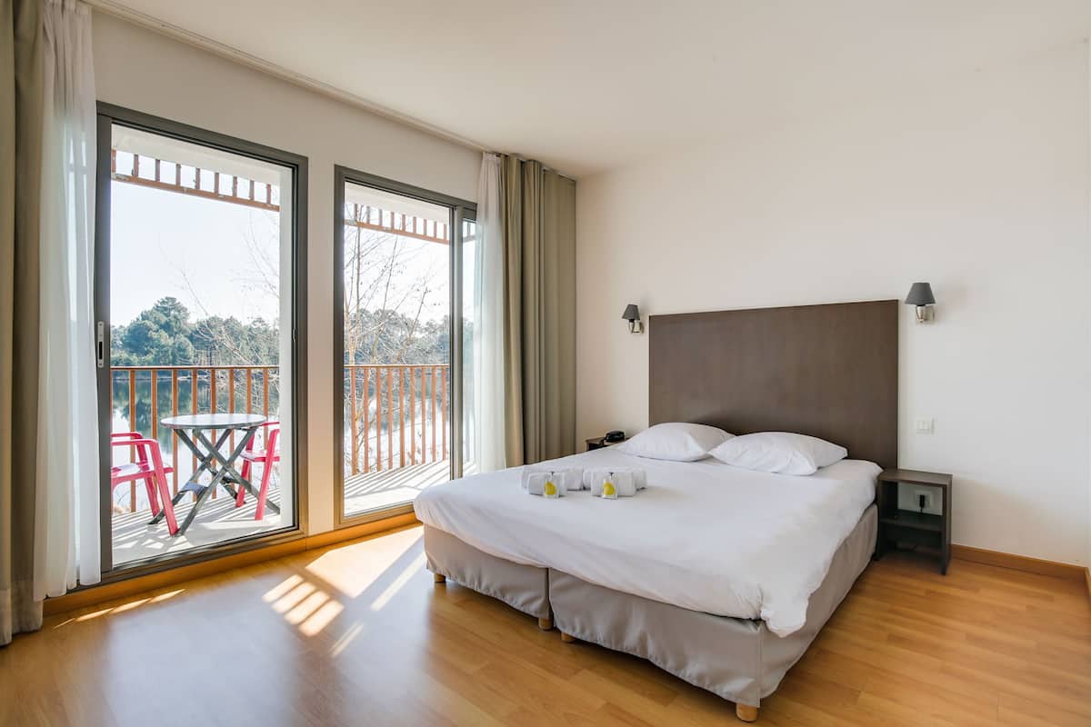 A hotel room with a large bed, a nightstand, and a view of a balcony with chairs and a table.