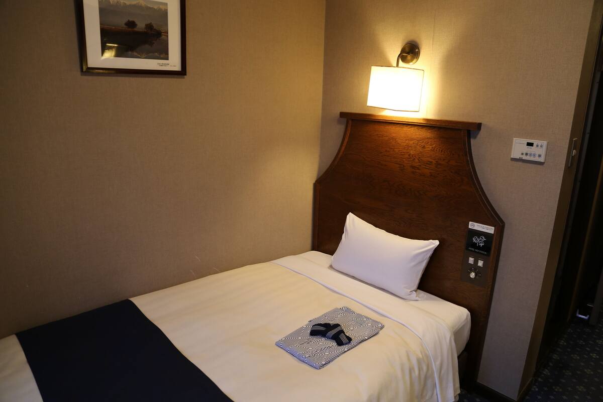 Habitación de hotel con una cama bien hecha, una lámpara de noche, un termostato y un cuadro en la pared.