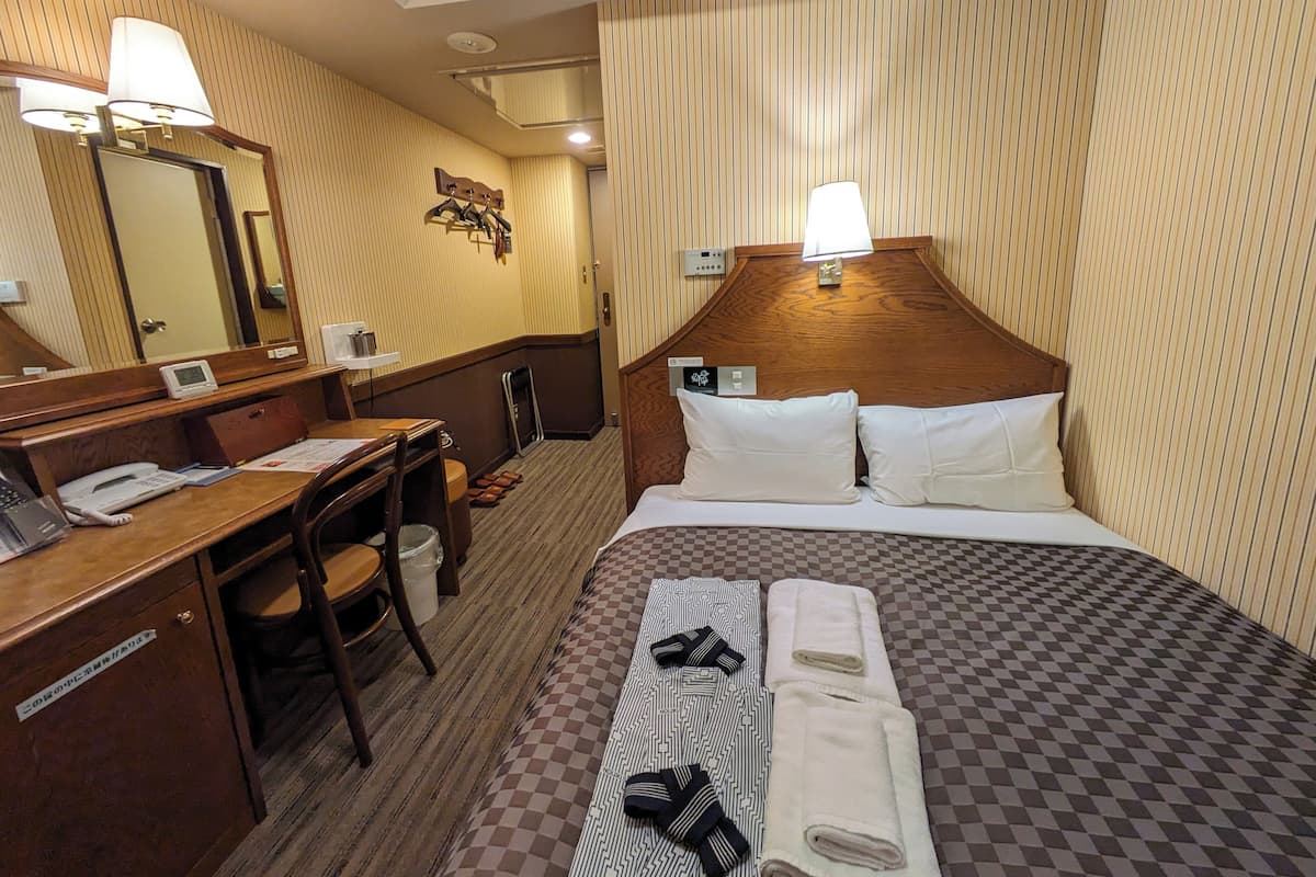 Habitación de hotel con cabecera de madera, un escritorio con teléfono, una silla y una cama con almohadas y una colcha a cuadros.