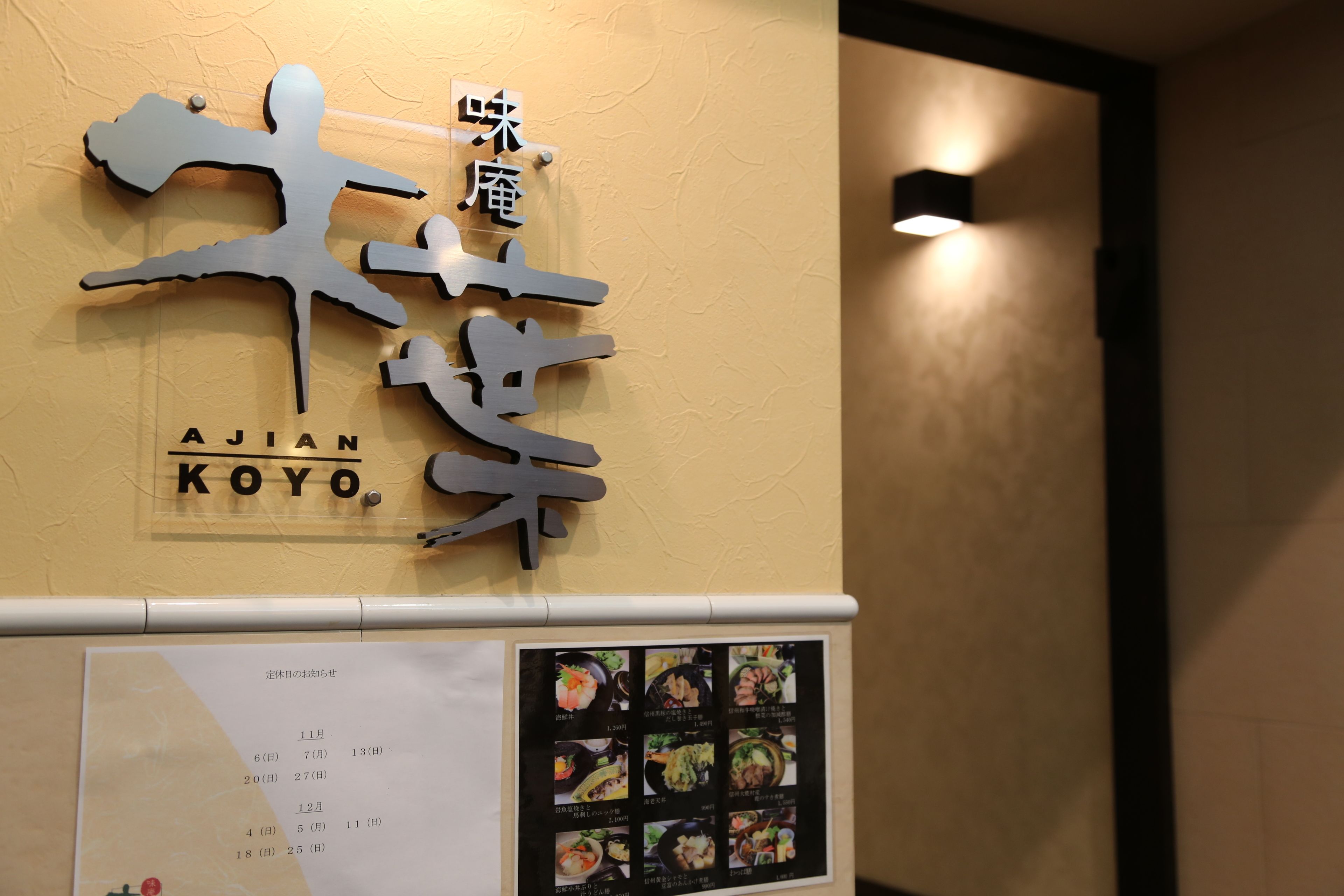 Un mur sur lequel est affiché un grand panneau indiquant « AJIAN KOYO », et en dessous, un tableau présentant le menu.