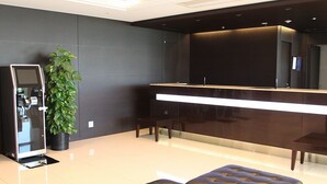 Uma área de recepção moderna com balcão, uma grande planta em vaso e um espaço de estar com dois pufes de couro preto.