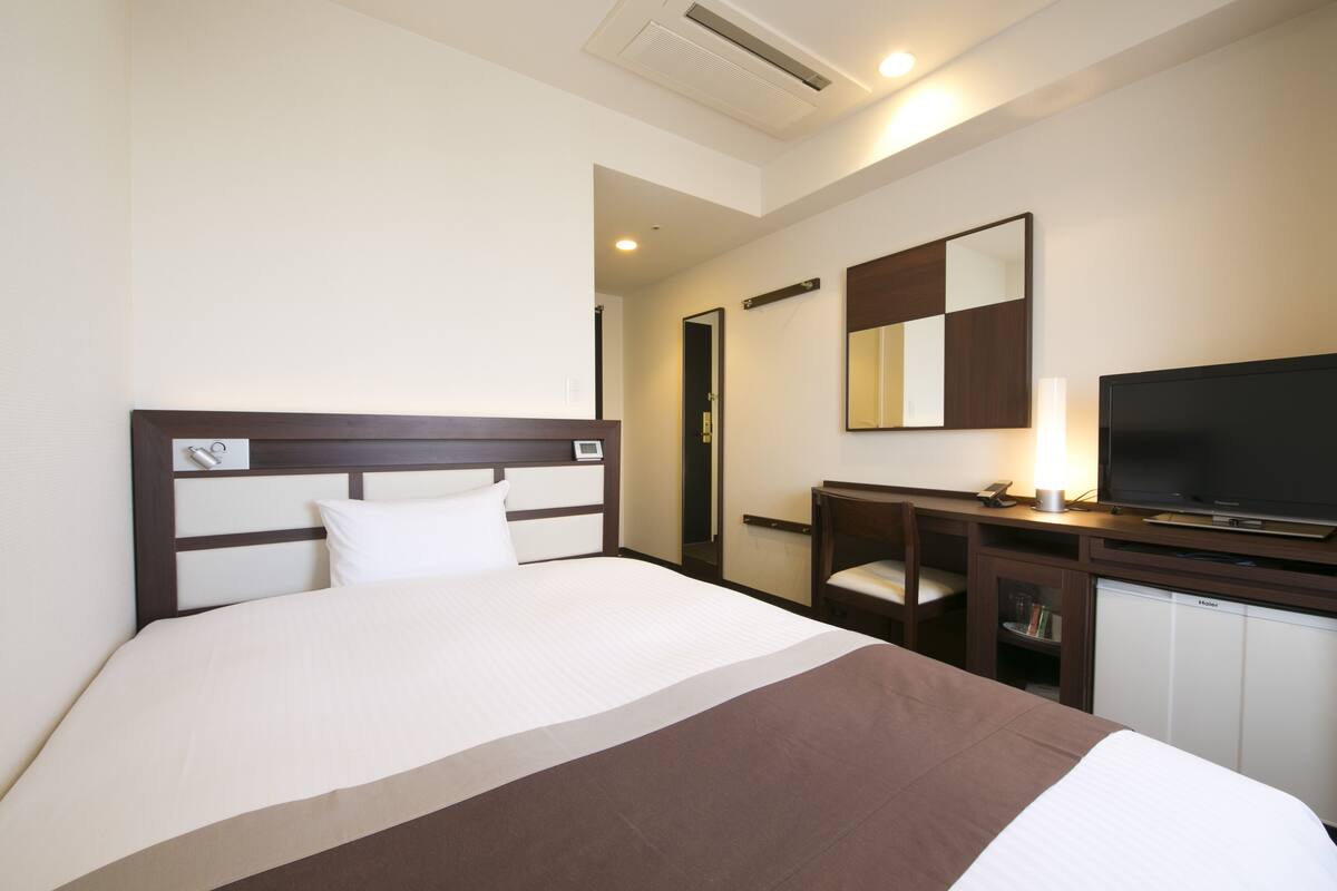 A hotel room with a large bed, a desk with a television, a mirror, and a minibar.