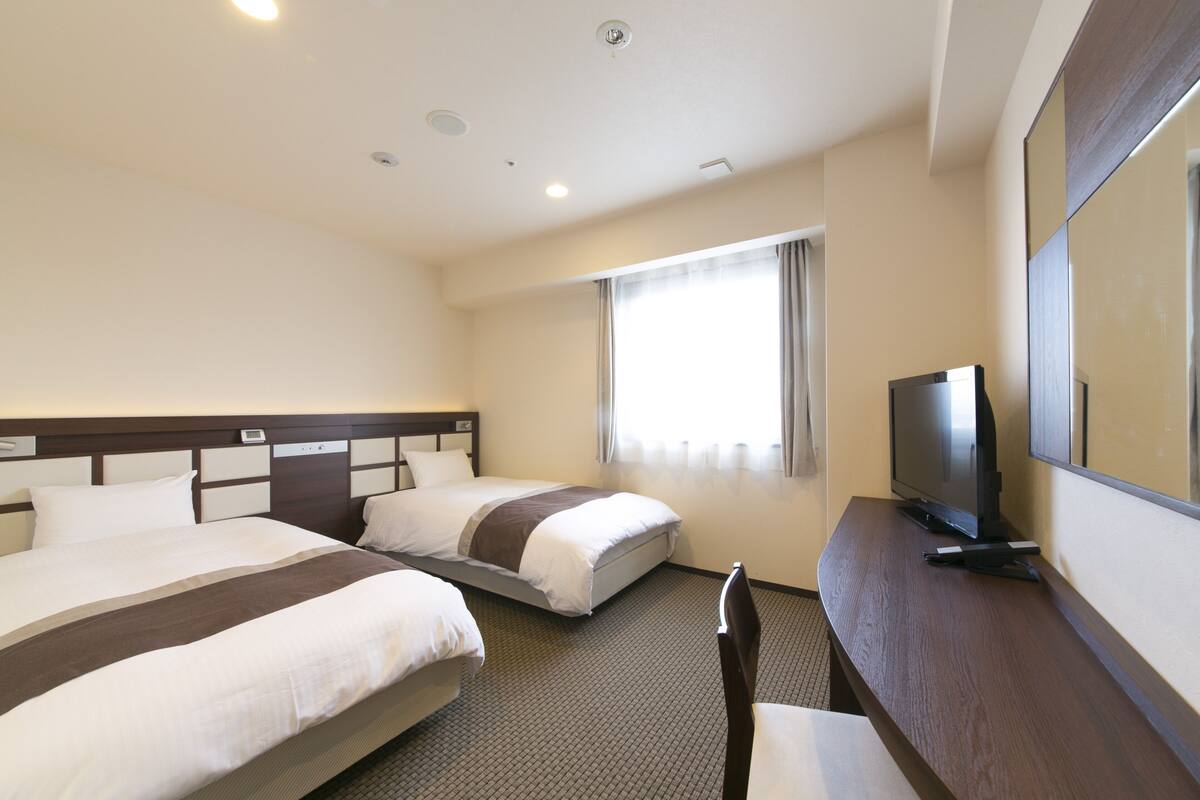 A hotel room with two beds, a desk with a TV, and a large window.