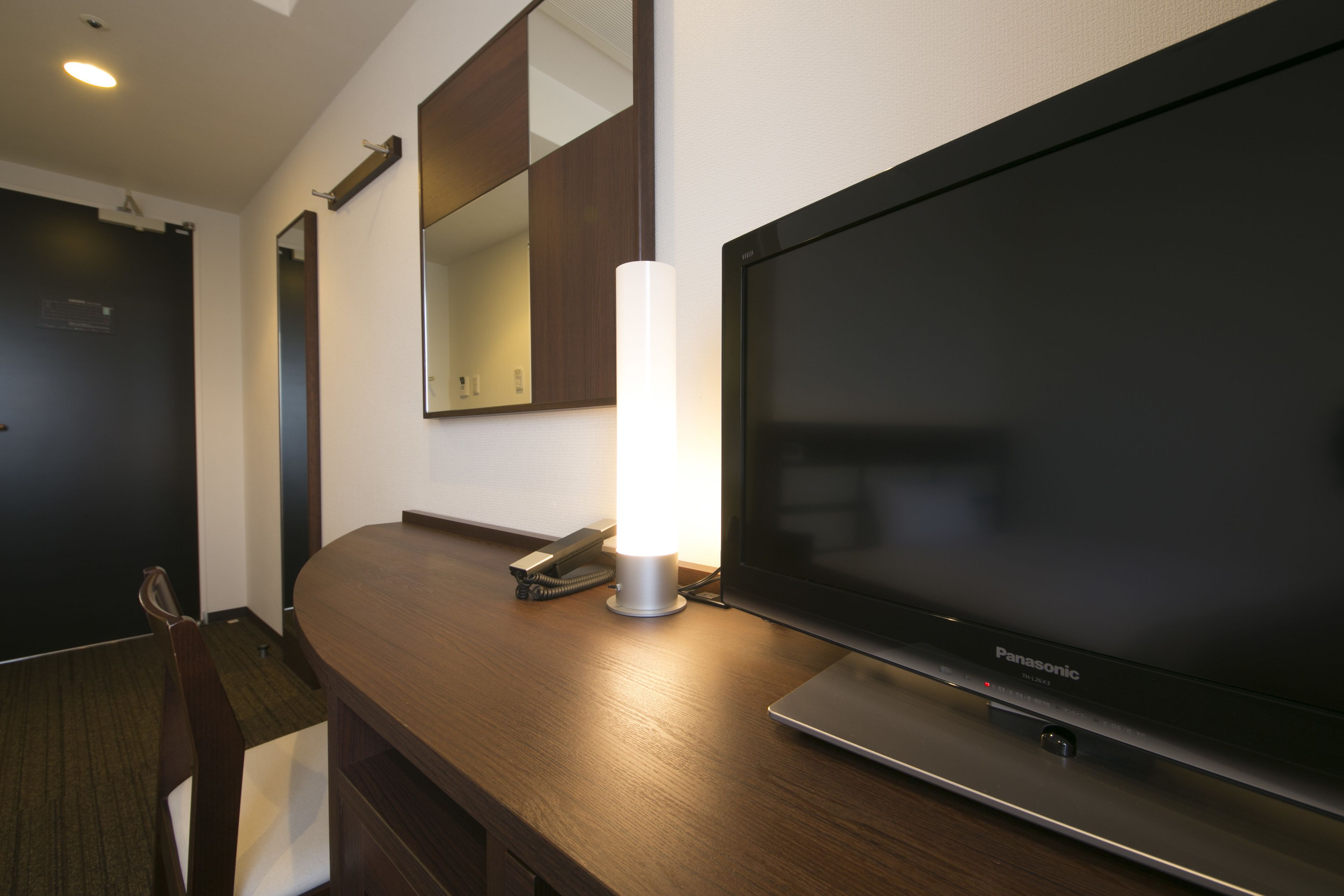 A modern hotel room with a large flat-screen TV, a bedside lamp, and a wooden desk.