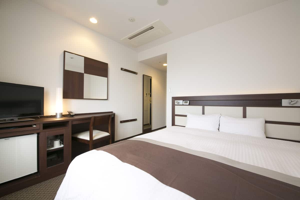A hotel room with a bed, a television, a desk with a chair, and a mirror on the wall.