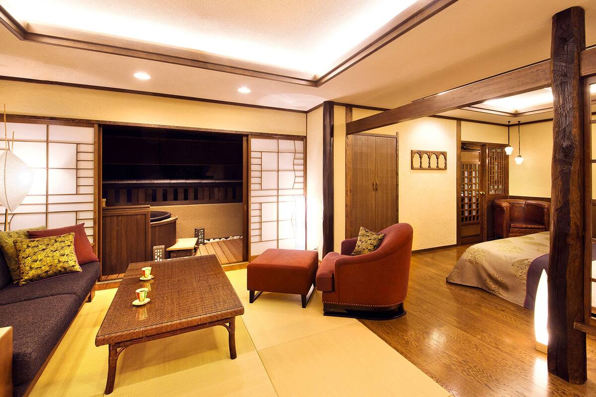 Une pièce de style japonais traditionnel, avec un sol recouvert de tatamis, une petite table avec des tasses, un canapé avec des coussins, un fauteuil rouge et un lit doté d’une tête de lit en bois.