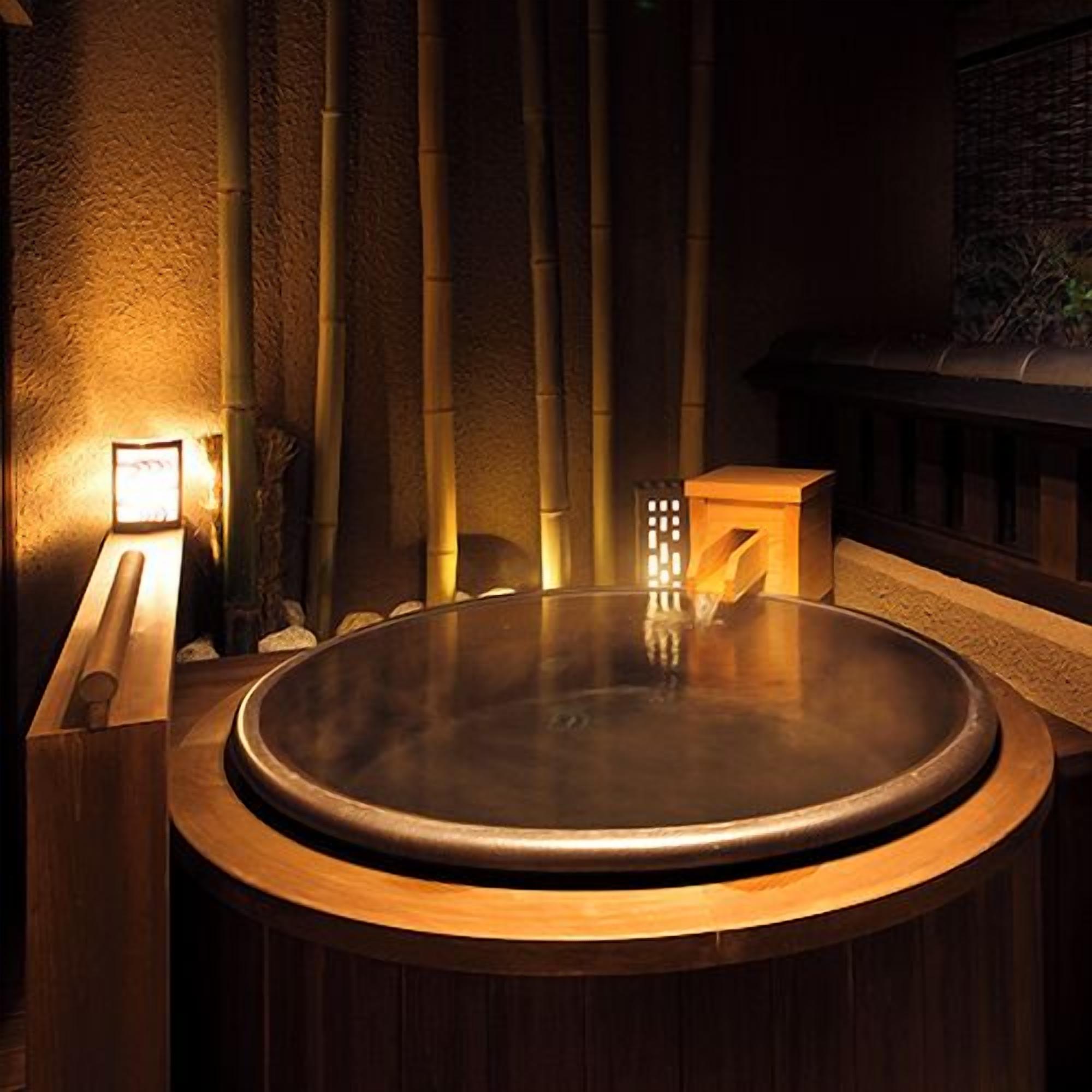 Un jacuzzi rond, doté d’un bord en bois, entouré de plantes de bambou et d’un mur de pierre.