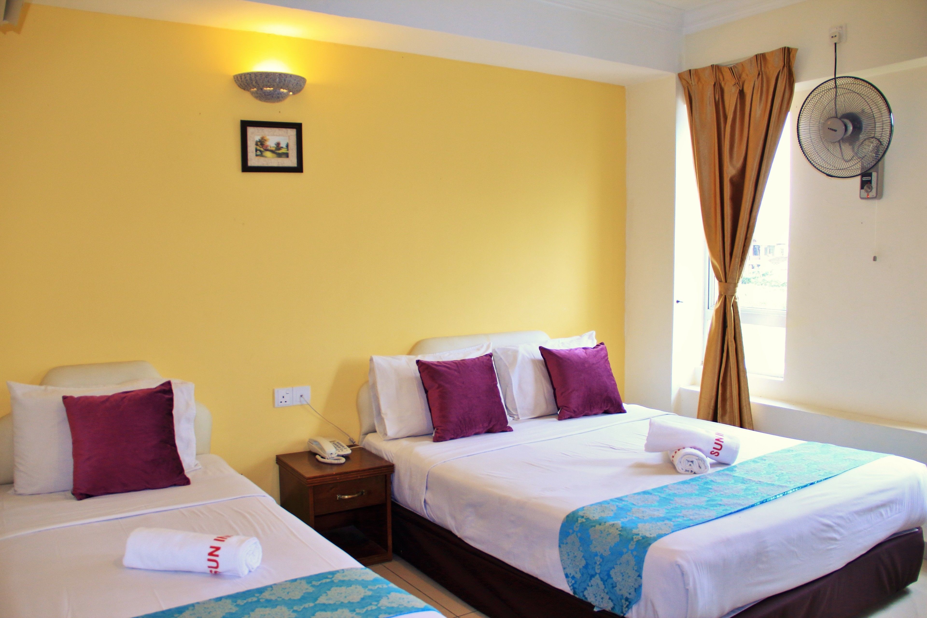 A hotel room with two beds, a nightstand with a phone, a wall-mounted fan, and a window with curtains.