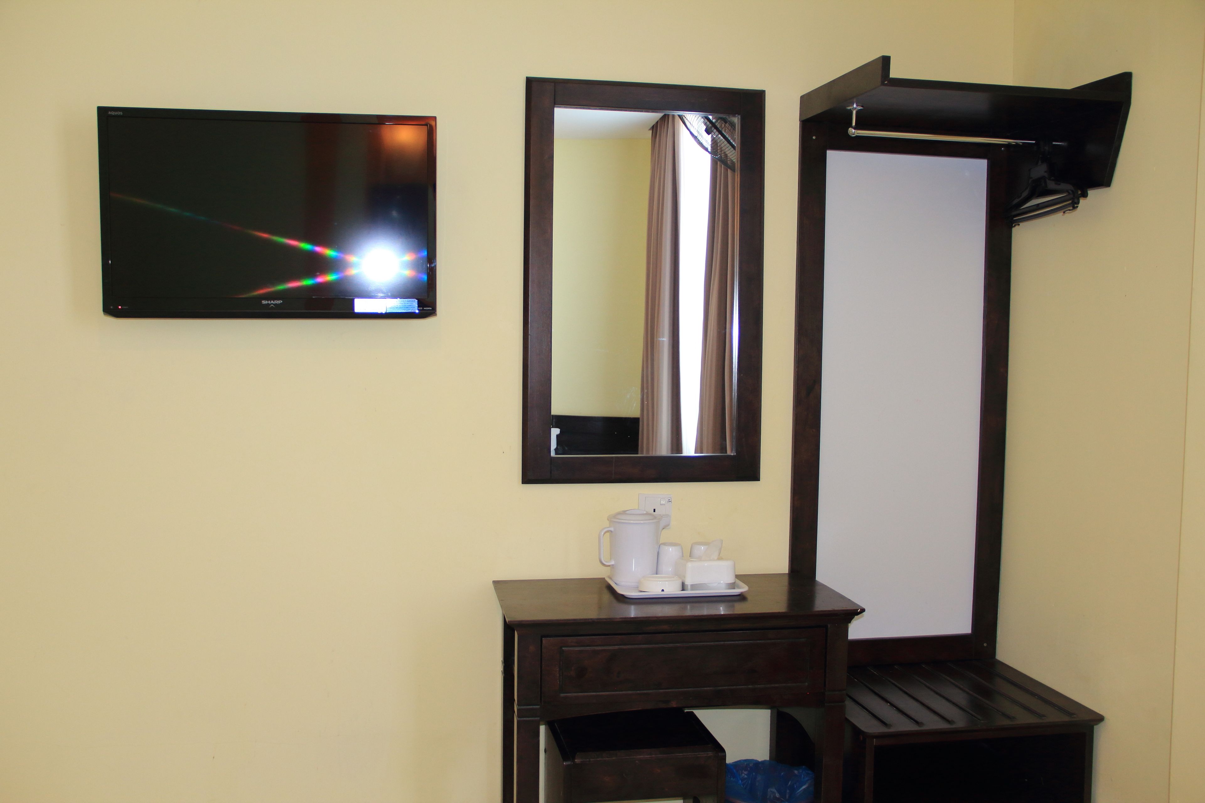 A hotel room with a flat-screen TV mounted on the wall, a mirror, a small wooden table with a coffee maker, and a trash bin.