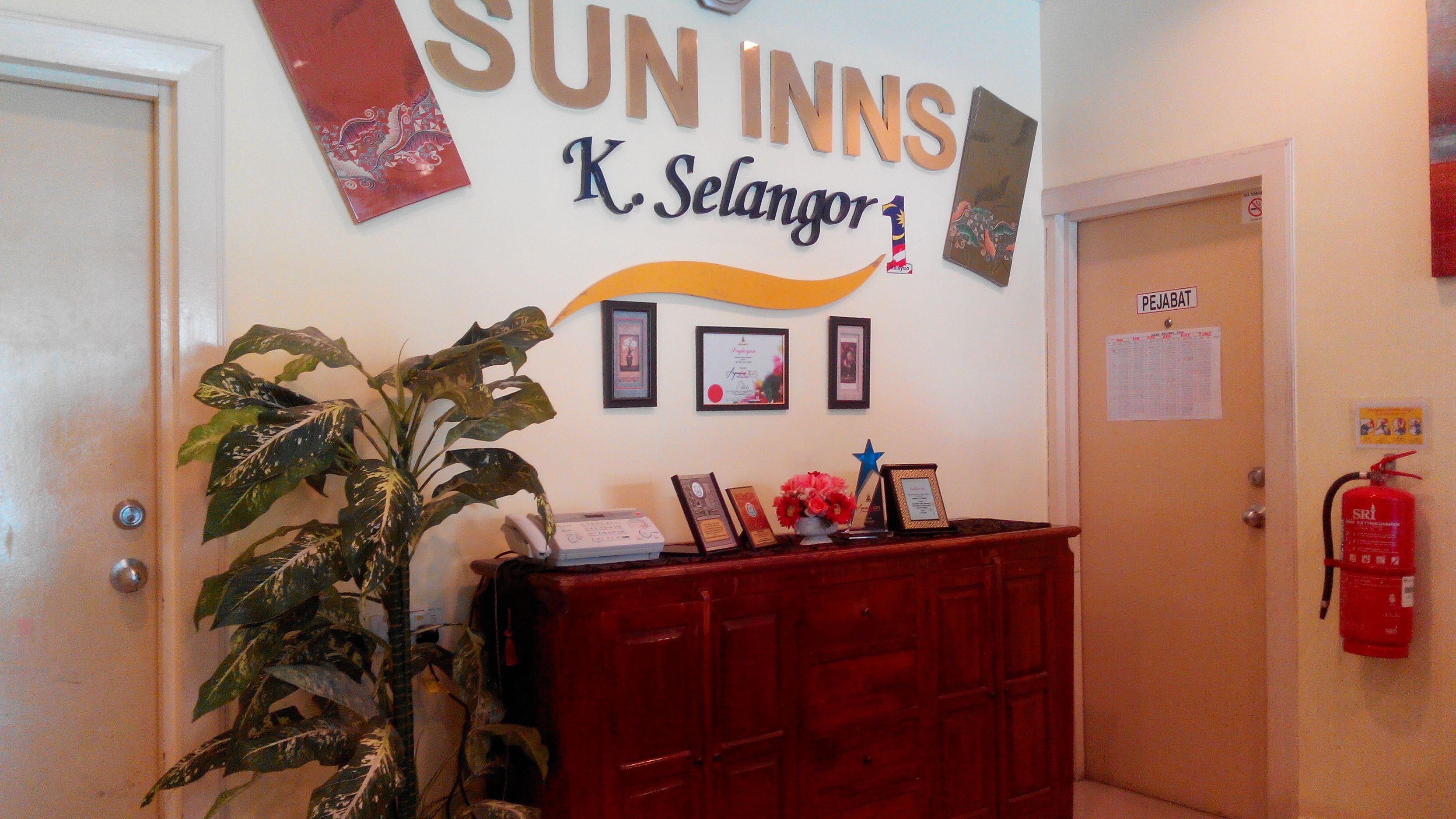 Uma área de recepção com uma placa indicando Sun Inns K. Selangor, uma mesa de madeira com certificados emoldurados, uma planta em vaso e um extintor de incêndio.