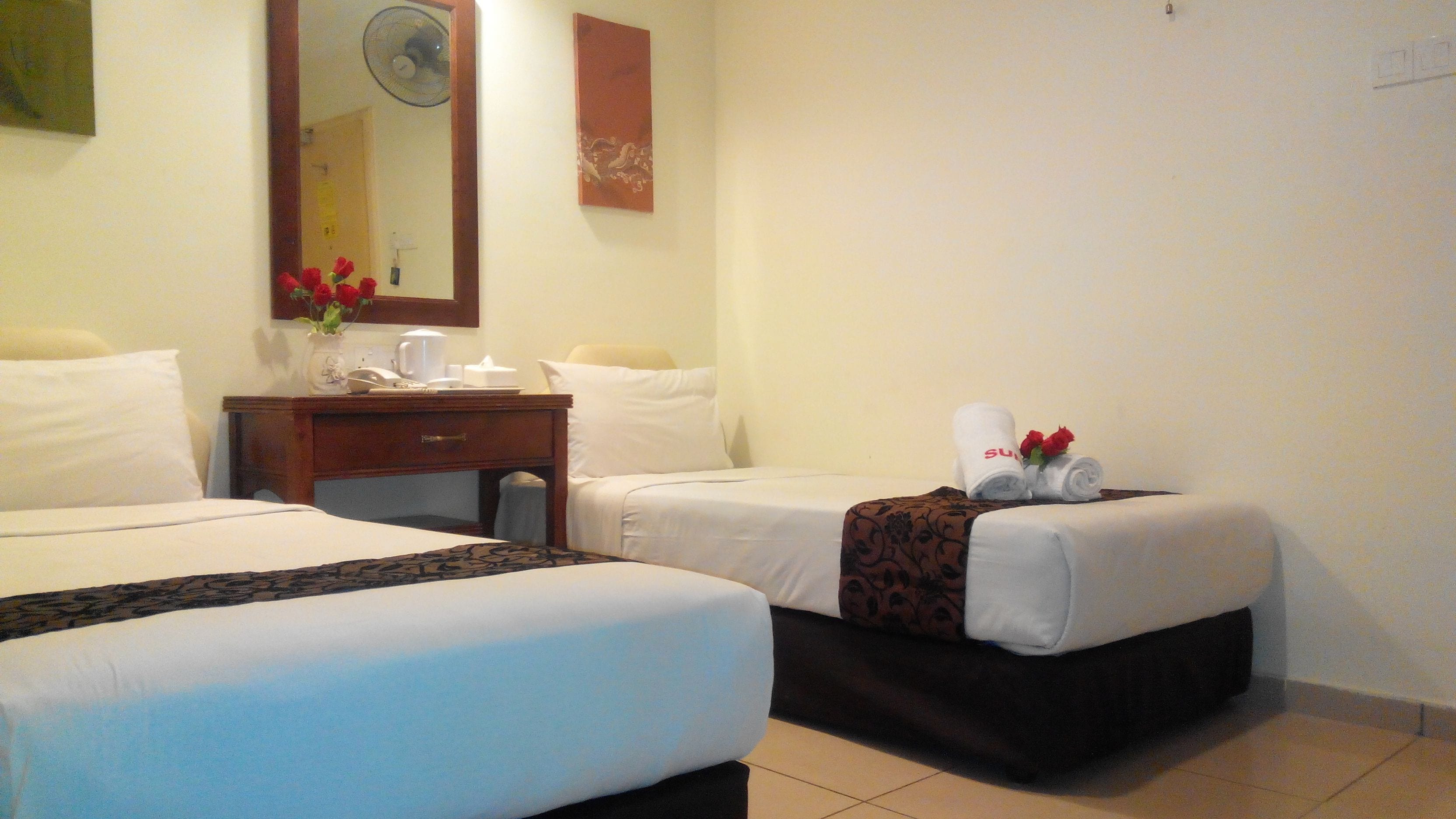 A hotel room with two beds, a wooden nightstand with a vase of red flowers, a mirror, and a wall-mounted fan.