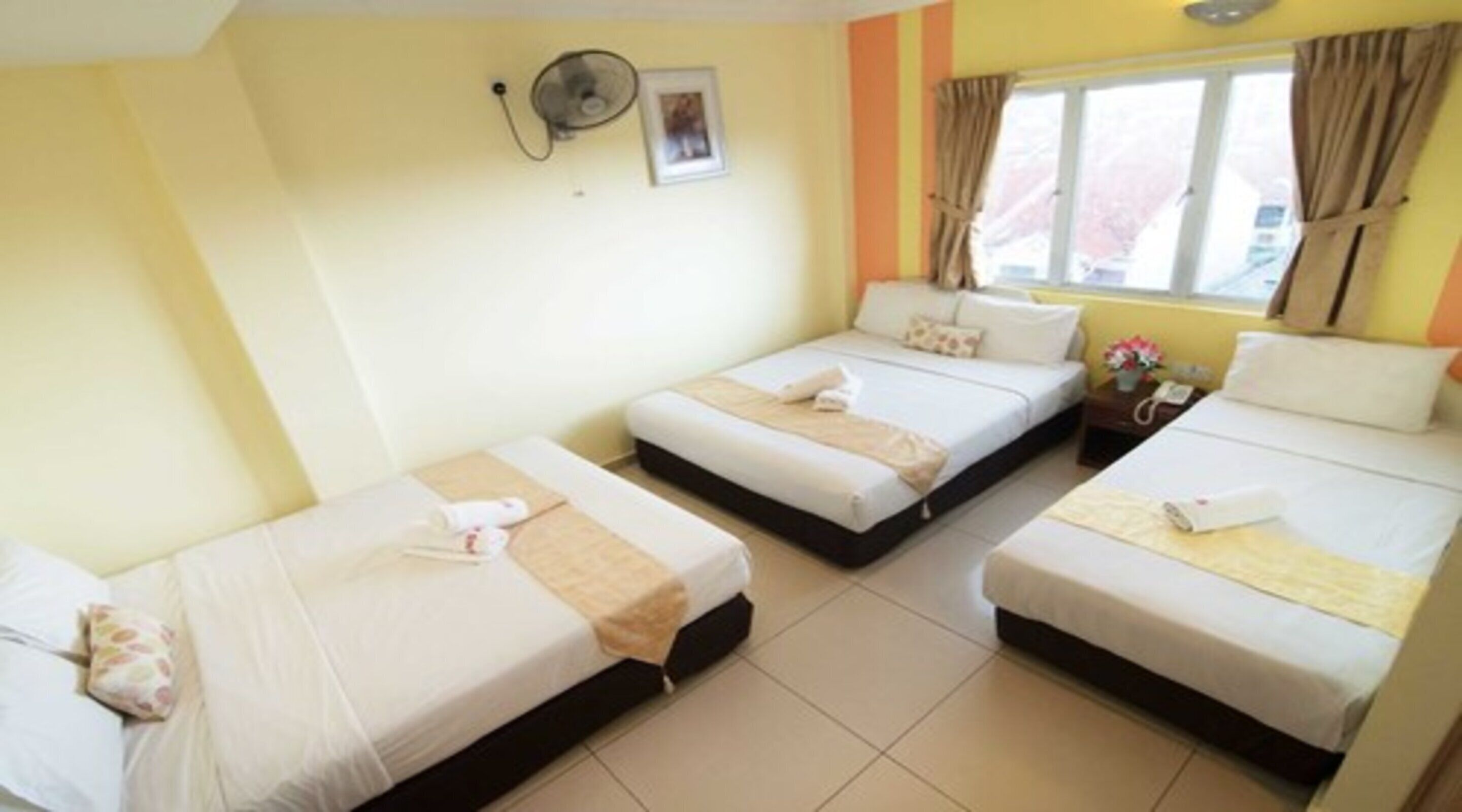 Una habitación de hotel con tres camas individuales, una mesita de noche con lámpara, una ventana con cortinas y un ventilador de pared.