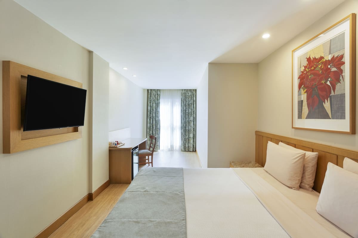 A hotel room with a bed, a desk, a chair, a television mounted on the wall, and a painting on the wall.