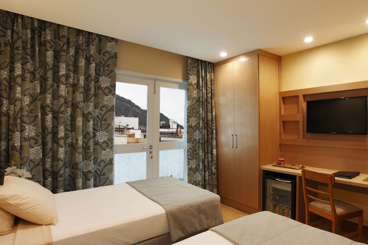 A hotel room with two beds, a TV, a desk, and a view of the sea.
