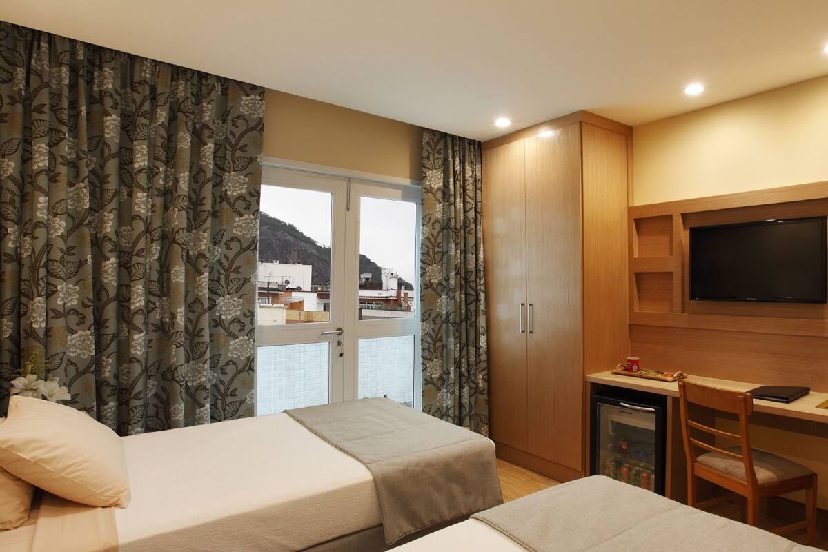 A hotel room with two beds, a TV, a desk, and a view of the sea.