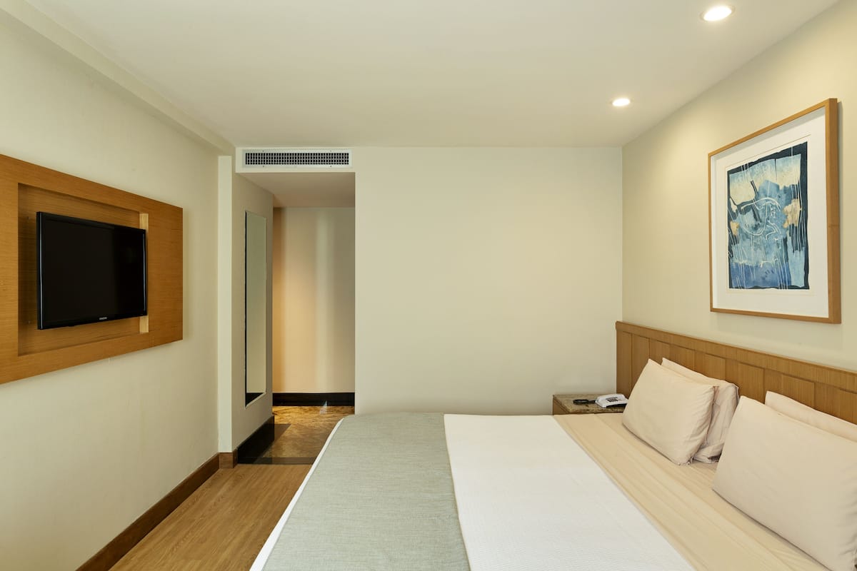 A hotel room with a large bed, a flat-screen TV mounted on the wall, a framed artwork above the bed, and a small bedside table with a phone.
