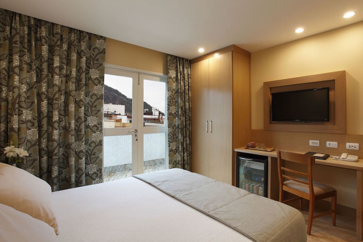 A hotel room with a bed, a desk with a chair, a television, and a view of a mountainous landscape through the window.