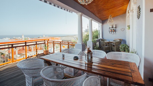 Terraza de madera con sillas de mimbre y una mesa, con vistas a un paisaje urbano.