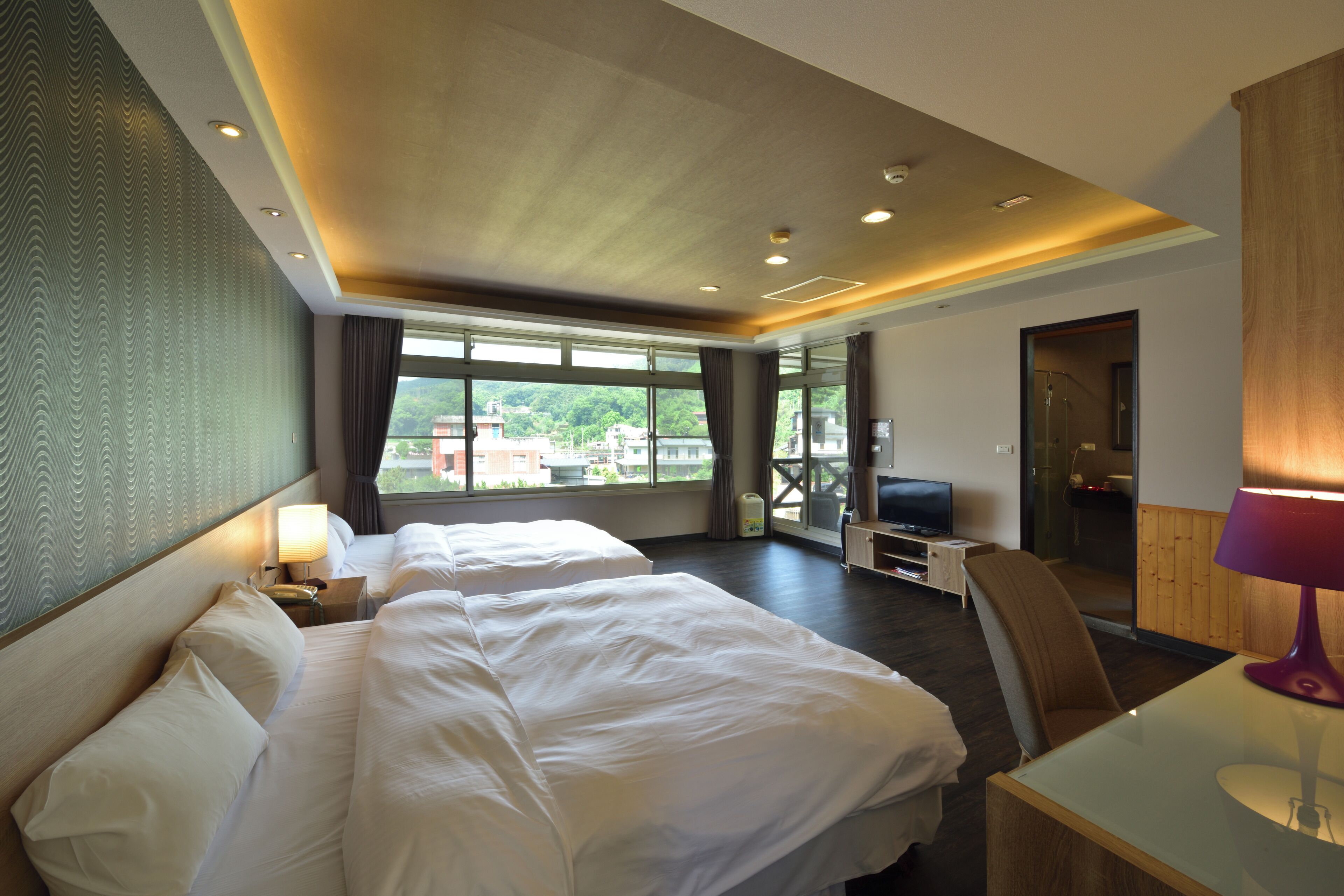 A hotel room with two beds, a desk, a TV, and a view of the city.