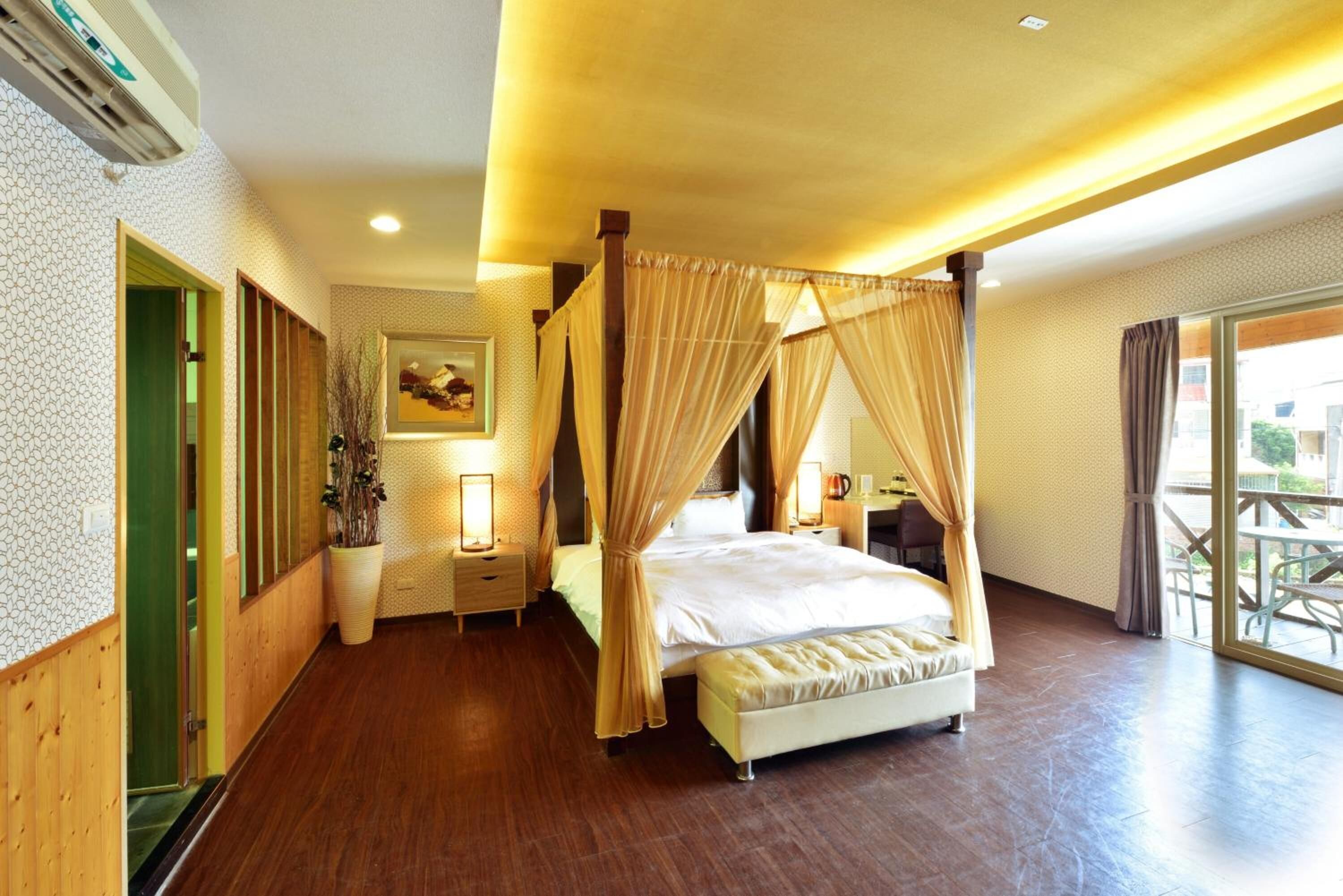 A spacious bedroom with a four-poster bed, a bedside table with a lamp, and a view of the outdoors through a large window.