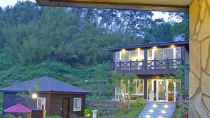 A wooden deck with two wicker stools and a glass table, overlooking a house with a stone wall and a balcony. - Cimengrou Hall (Dahu)
